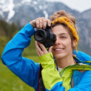 delighted-young-european-woman-takes-photo-during-hiking-trip-holds-professional-camera
